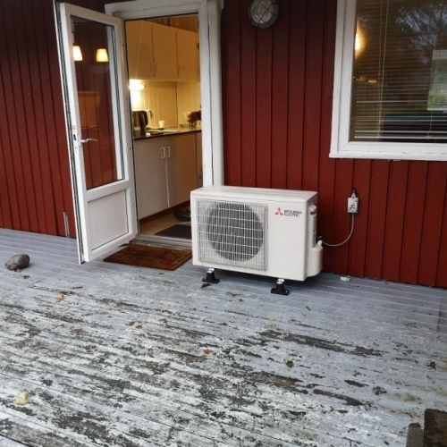 Udendørs varmepumpe på terrasse ved rødt træhus.