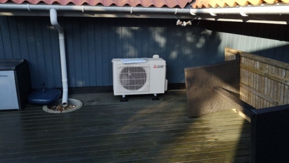 Udendørs varmepumpe på terrasse ved mørkt træhus.