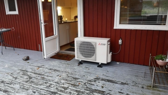 Udendørs varmepumpe på terrasse ved rødt træhus.